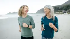 Sahilde koşan 50lerinde iki kadıni biri kıvırcık biri düz sarı saçlı, üzerilerinde ince body ve tayt var. Sahilde onlardan başka kimse yok ve arkaları dağlık.