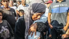 Uma mulher chora segurando um bebê no colo. Várias pessoas ao redor olham