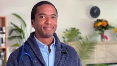 Un hombre con una chaqueta azul y una camisa celeste y un estetoscopio sonríe en una sala de estar.