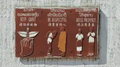 Placa na parede com orientações aos turistas sobre como devem se comportar, escritas em laociano, inglês e chinês, além de ilustrações