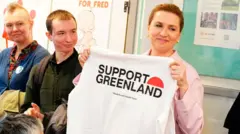 Улыбающаяся Метте Фредериксен стоит на фоне стенда, в руках у нее футболка с надписью Support Greenland.