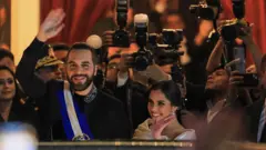 nayib bukele rodeado de fotógrafos. levanta la mano y saluda. sonríe y tiene puesta la banda presidencial.