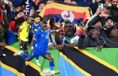 Umukinyi atera umupira, inyuma abafana bafise amadarapo ya Tanzania na Uganda