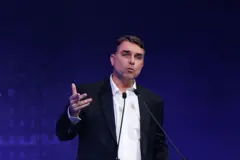 Flávio Bolsonaro aparece de blazer escuro e camisa branca, durante discurso, com um fundo azul