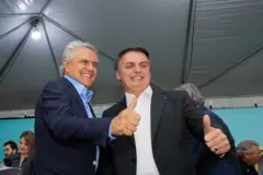 Caiado e Bolsonaro sorrindo para foto e fazendo joinha; eles estão embaixo de tenda de evento, com outras pessoas à volta
