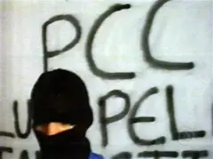 Uma pessoa com o rosto coberto em frente a uma parede em que se lê PCC.