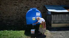 Uma mulher idosa enche um balde com água de um reservatório instalado na rua na vila de Gorna Studena, na Bulgária.