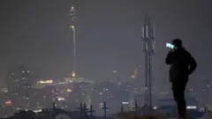 Um homem está em pé no alto de uma colina, segurando um celular junto ao ouvido, com a tela iluminada. Ao longe, aparecem as luzes de Teerã à noite, com uma grande torre de telecomunicações e, mais distante, uma antena de telefonia móvel