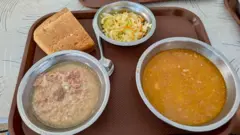 Três tigelas metálicas sobre uma bandeja com um pedaço de pão, duas delas com sopa, uma com carne, e salada na terceira