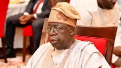 Nigeria President, Bola Tinubu wear cream colour cloth wit brown designs e also wear cream colour cap with broun design and eye glasses as e siddon for wooden chair with red design.