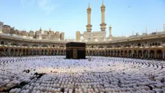 Десятки мусульман в белом стоят на коленях и молятся вокруг черного куба в Мекке.
