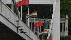Kínai és magyar zászlók sorakoznak egy hídon Budapesten, a háttérben egy szobor és oszlopos építmény látható a Gellért-hegy alján.