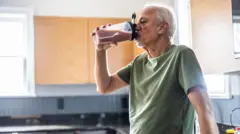 Un hombre con camiseta verde bebe un batido de una botella de plástico. Está apoyado en un costado de la cocina.