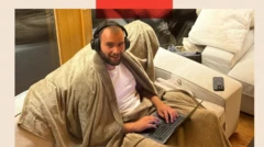 Мужчина в найшниках, закутавшись в плед, сидит на диване с ноутбуком на коленях и с улыбкой смотрит в камеру