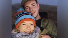 A médica Caroline Muirhead, usando um gorro de lã azul e vermelho e um cachecol cinza, com Alexander “Sandy” McKellar ao fundo, de boné e casaco escuro