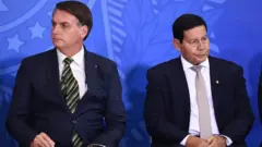Bolsonaro e Mourão sentados, olhando para lados opostos, em evento
