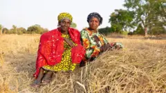 Deux femmes assises sur l'herbe jaune et sèche d'un champ, portant des foulards, des robes et des sandales colorées ; plusieurs arbres et arbustes sont visibles en arrière-plan.