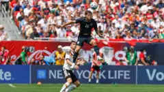 黒っぽいユニホームの10番の選手が空中高くジャンプしてヘディングしている。すぐ後ろには白のユニホームの選手がいる