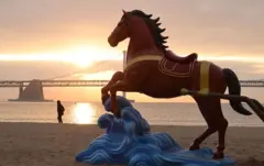 모래사장에 파도 위를 달리는 붉은 말 조각상이 설치돼 있다. 뒷 배경으로는 바다를 가로지르는 다리가 보인다.