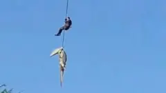 Une capture d'écran granuleuse montrant une corde au bout de laquelle se trouve un crocodile, tandis qu'un homme s'y accroche plus haut