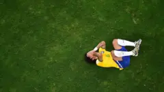 A imagem mostra um jogador da seleção brasileira de futebol caído no gramado, encolhido e com as mãos cobrindo o rosto, sugerindo dor ou frustração após um lance. Registrada de um ângulo aéreo, a fotografia destaca o contraste entre a figura do atleta e a ampla área verde do campo, reforçando a sensação de isolamento no momento.