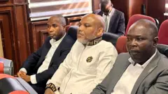Nnamdi Kanu wia white Fendi siddon in-between to security officers