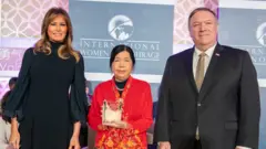 Susanna Liew veste um casaco bordado em vermelho e amarelo e ergue um troféu de vidro, com um leve sorriso, enquanto está no palco entre Melania Trump, que usa um vestido preto, e Mike Pompeo, que veste um terno preto e gravata dourada