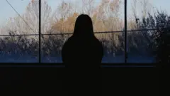 La silueta de una mujer mirando por una ventana, con árboles y follaje al fondo.