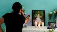 Mãe de América Sánchez de costas, voltada para um altar improvisado com velas, flores e fotos da filha