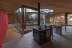 Interior de una casa con grandes ventanales que dan a un patio arbolado, una cocina con estufa antigua y una mesa de madera bajo un techo inclinado revestido en madera.