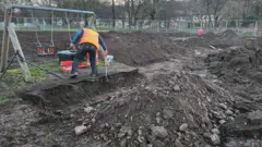 Terra revirada no parquinho com um balanço no meio, e um homem cavando em frente a ele