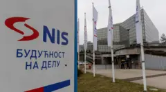 Velika tabla na kojoj je napisano plavim slovima NIS, crveni znak firme pored, a ispod ćirilično - Budućnost na delu. U daljini je staklena zgrada kompanije. 