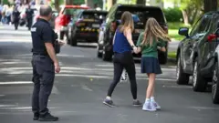 Un policía en medio de la calle supervisa mientras dos chicas avanzan de la mano. Todos aparecen de espaldas.