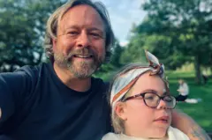 Darren y Sophia Scott. Él tiene el cabello rubio, algo largo, y barba, y lleva puesta una camiseta azul. Tiene el brazo alrededor de Sophia, quien lleva gafas y una bufanda de algodón azul, marrón y blanca atada alrededor de la cabeza. Él sonríe a la cámara, mientras que ella mira hacia fuera de cuadro. Se encuentran en un parque.