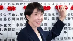 Санаэ Такаити - женщина в темном пиджаке - на фоне плаката с японскими иероглифами