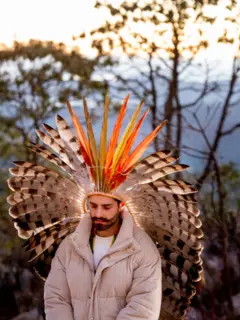 Alok de olhos fechados em meio à natureza, usando um cocar de grandes penas em tons de branco, preto, laranja e amarelo, que ocupa quase toda a composição. Vestido com um casaco acolchoado bege, ele aparece em uma postura contemplativa.