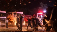 Çıkan orman yangınına müdahale edenlerin toplanma alanındaki görüntüsü, tırlar, ellerini havaya kaldırmış kişiler, koşan görevliler ve arkada yanan orman var. Gece görüntüsü