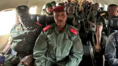 Brigadier General Olivier Gasita (hagati imbere) hamwe n'abandi basirikare bakuru muri FARDC bicaye bari mu ndege mu gihe cyashize. Gasita arareba muri 'camera', yambaye imyenda ya gisirikare y'icyatsi kibisi iriho amapeti atukura ku ntugu n'ingofero y'umutuku wijimye. 