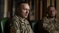 Близько 40-річний чоловік у військовій формі Збройних сил України сидить за столом під час наради. На рукаві видно український прапор і шеврон із тризубом. На задньому плані, розмитий, сидить Павло Паліса.