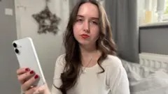 Fenella está segurando seu celular substituto na sala de estar. Ela está triste. O ambiente ao seu redor é decorado em tons suaves de cinza e marfim. Ela tem cabelos castanhos compridos e olhos castanhos.