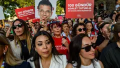 Çok sayıda kadın, protestoda yan yana ayakta duruyor. Ellerindeki pankartlarda "100 gündür adalet tutuklu, biz özgürlüğün peşindeyiz" yazıyor. Ortadaki bir kadın elinde Ekrem İmamoğlu'nun maskesini kaldırıyor ve kadının tişörtünde "İmamoğlu her yerde" yazıyor