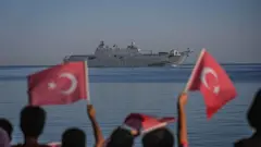 Çocukların flu bayran sallayan görüntüleri önde, hepsinin arkası dönük denizden geçen gemiye bakıyorlar. 