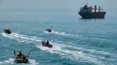 Cuatro lanchas rápidas con banderas iraníes navegando por el Golfo Pérsico, cerca de un buque de carga general.