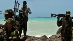 20 Ağustos 2010'da Somali'nin merkezindeki Hobyo kasabasının kayalık bir çıkıntısında silahlı milis üyeleri ve bir korsan yürürken, ufukta kaçırılmış bir süper tanker demirlemiş halde görülüyor.