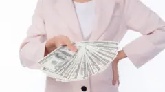 سيدة تمسك دولارات. ترتدي جاكيت زهري بأزرار سوداء وقميص أبيض، لا يظهر رأسها في الصورة.