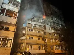 Пострадавшее жилое здание, в котором видно летающие окна и разрушенные потолки. На верхних этажах здания находится огонь.