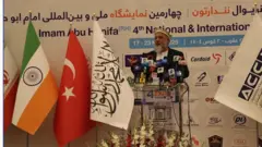 وزیر تجارت حکومت طالبان با پیراهن سفید، کرتی سیاه و کلاه روشن پشت میز سخنرانی در یک نمایشگاه که پرچم چند کشور در کنارش قرار دارد