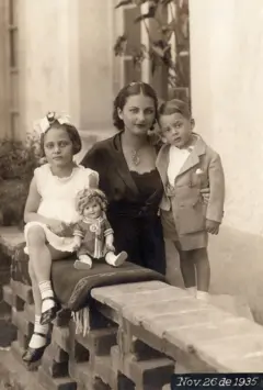 Cristina, no centro, usa um vestido e uma blusa pretos e um colar. Ao seu lado está Antonio, que usa uma camisa branca, uma jaqueta e um short cinza. Do outro lado, sentada, está Isolda, que veste um traje branco sem mangas, meias brancas, sapatos pretos e um laço na cabeça. Uma boneca está sentada à frente delas. Estão em um jardim