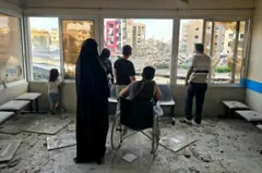 Siyah çarşaflı bir kadın, tekerlekli sandalyede bir erkek ve önlerinde üç kişi ve bir kız çocuğu yıkık bir bekleme salonunun camından yıkıntılarla dolu bir alana bakıyor, hepsinin arkası dönük. 