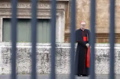 Cardeal católico de pé atrás de uma grade, em frente a um santuário